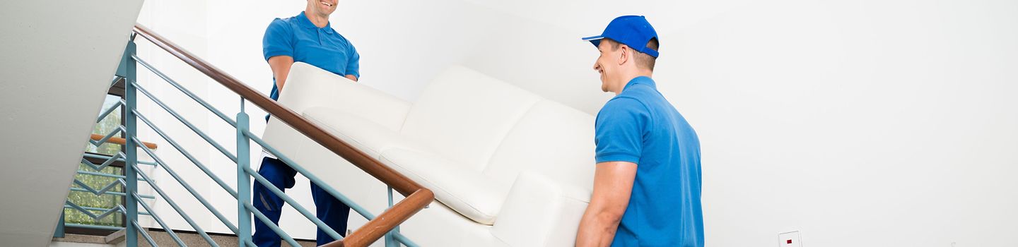 Zwei Männer in blauer Arbeitskleidung tragen gemeinsam ein Sofa eine Treppe hinauf.