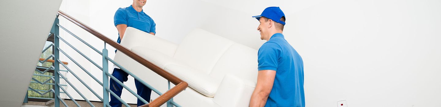 Zwei Männer in blauer Arbeitskleidung tragen gemeinsam ein Sofa eine Treppe hinauf. Zwei Männer in blauer Arbeitskleidung tragen gemeinsam ein Sofa eine Treppe hinauf.