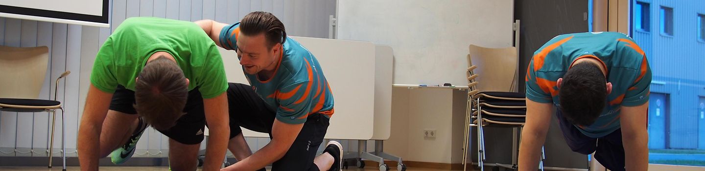 In einer Sporthalle befinden sich zwei Männer in Stützhaltung auf Sportmatten. Ein Mann macht unter Anleitung eines Sporttrainers eine Übung auf der Gymnastikmatte