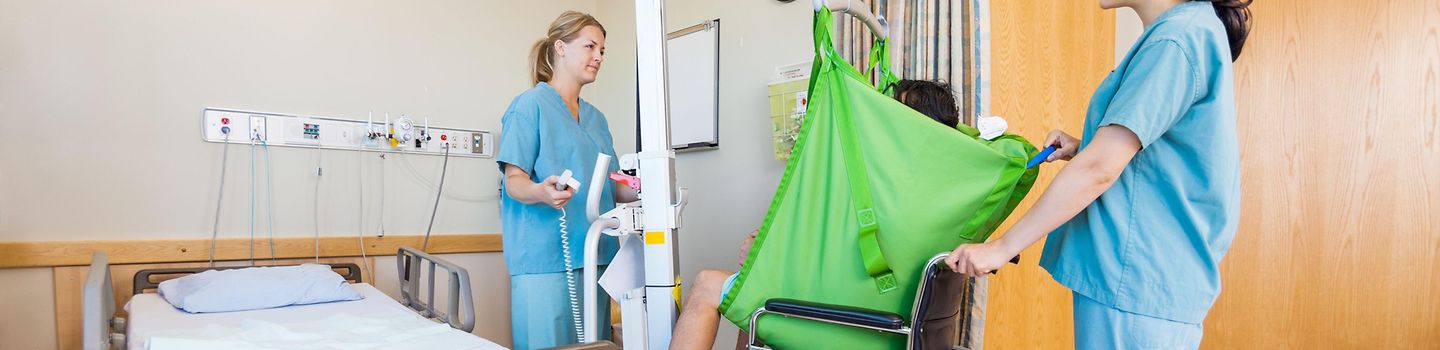 In einem Krankenhauszimmer benutzen zwei Krankenschwestern eine Hebehilfe, um den Patienten in einen Rollstuhl zu setzen