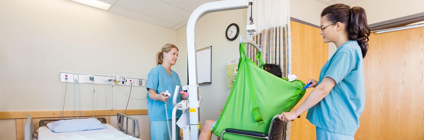 In einem Krankenhauszimmer benutzen zwei Krankenschwestern eine Hebehilfe, um den Patienten in einen Rollstuhl zu setzen In einem Krankenhauszimmer benutzen zwei Krankenschwestern eine Hebehilfe, um den Patienten in einen Rollstuhl zu setzen
