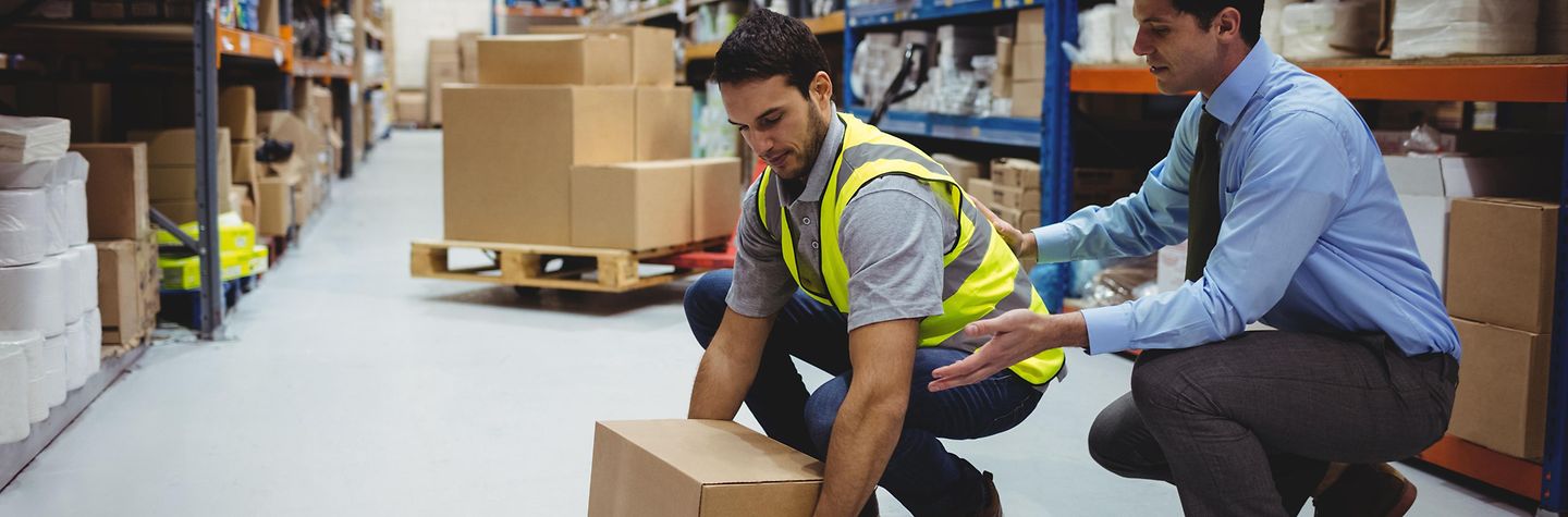 Ein Mann zeigt einem Lagerarbeiter wie ein Paket rückenschonend gehoben wird. Ein Mann zeigt einem Lagerarbeiter wie ein Paket rückenschonend gehoben wird