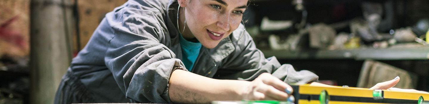 Eine Frau in einer Werkstatt bückt sich über die Werkbank und greift zur Wasserwaage.