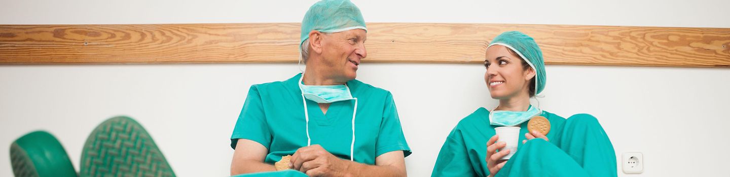 Ein Mann und eine Frau in OP-Kleidung sitzen auf dem Fußboden und unterhalten sich. Ein Mann und eine Frau in OP-Kleidung sitzen auf dem Fußboden und unterhalten sich.