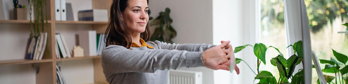 Homeoffice Junge Frau arbeitet zu Hause an einem Notebook. Sie streckt die Arme nach vorne und macht Dehnungsübungen.
