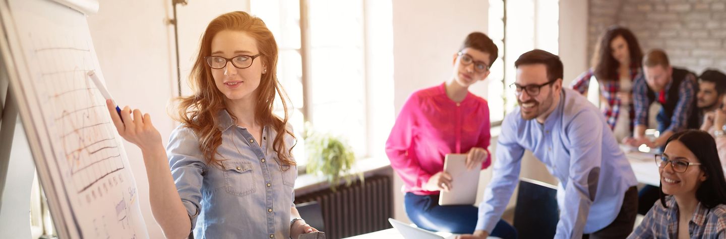 Ein Workshop oder Seminar wird dargestellt. Der Bildausschnitt zeigt zwei Reihen Tische. Junge Menschen sitzen und stehen an Tischen. Sie lachen miteinander und schauen auch die Frau an, die vorne steht. Sie zeigt mit einem Stift auf eine Stelle eines beschriebenen Flipcharts.